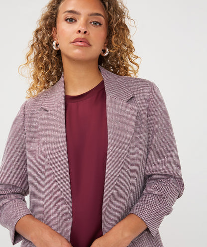 A woman with curly blonde hair wearing a textured, fine-checkered lavender and gray blazer over a deep burgundy top, photographed against a clean white studio background.