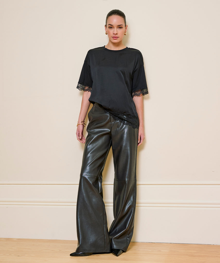 A model stands against a cream-colored wall wearing a black short-sleeve top with lace trim at the cuffs and black wide-leg faux leather pants.