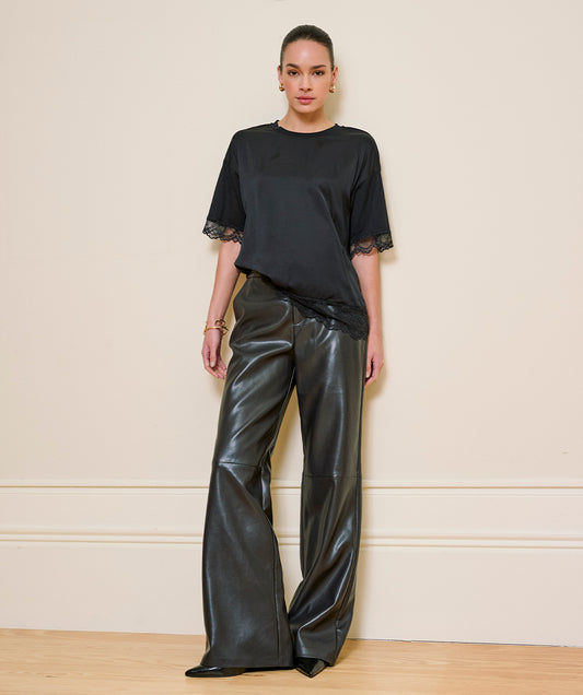 A model stands against a cream-colored wall wearing a black short-sleeve top with lace trim at the cuffs and black wide-leg faux leather pants.