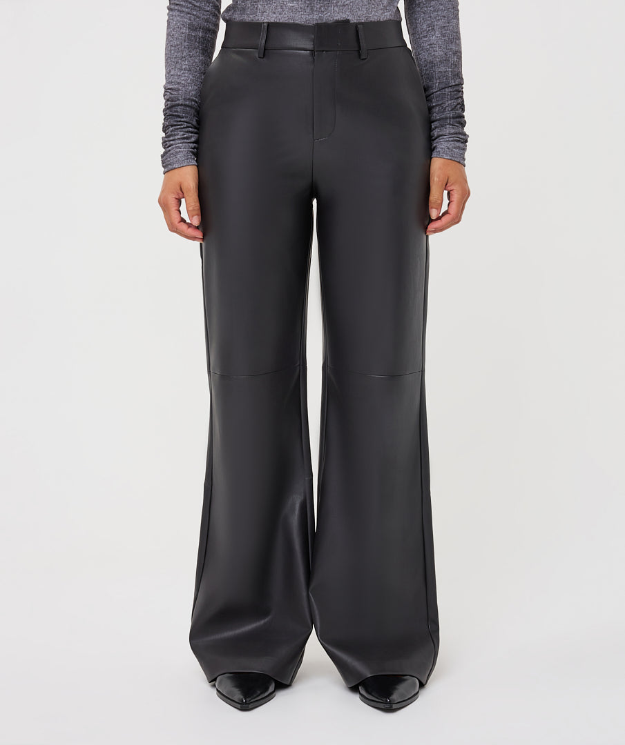 A model is shown from the waist down wearing high-waisted, wide-leg black faux-leather trousers, a gray long-sleeve top, and black pointed-toe heels against a light gray studio background.