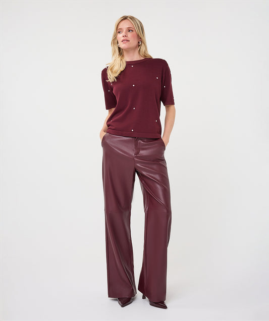 A woman modeling a short-sleeve burgundy knit top with small silver embellishments and matching wide-leg faux-leather pants against a plain light gray studio background.