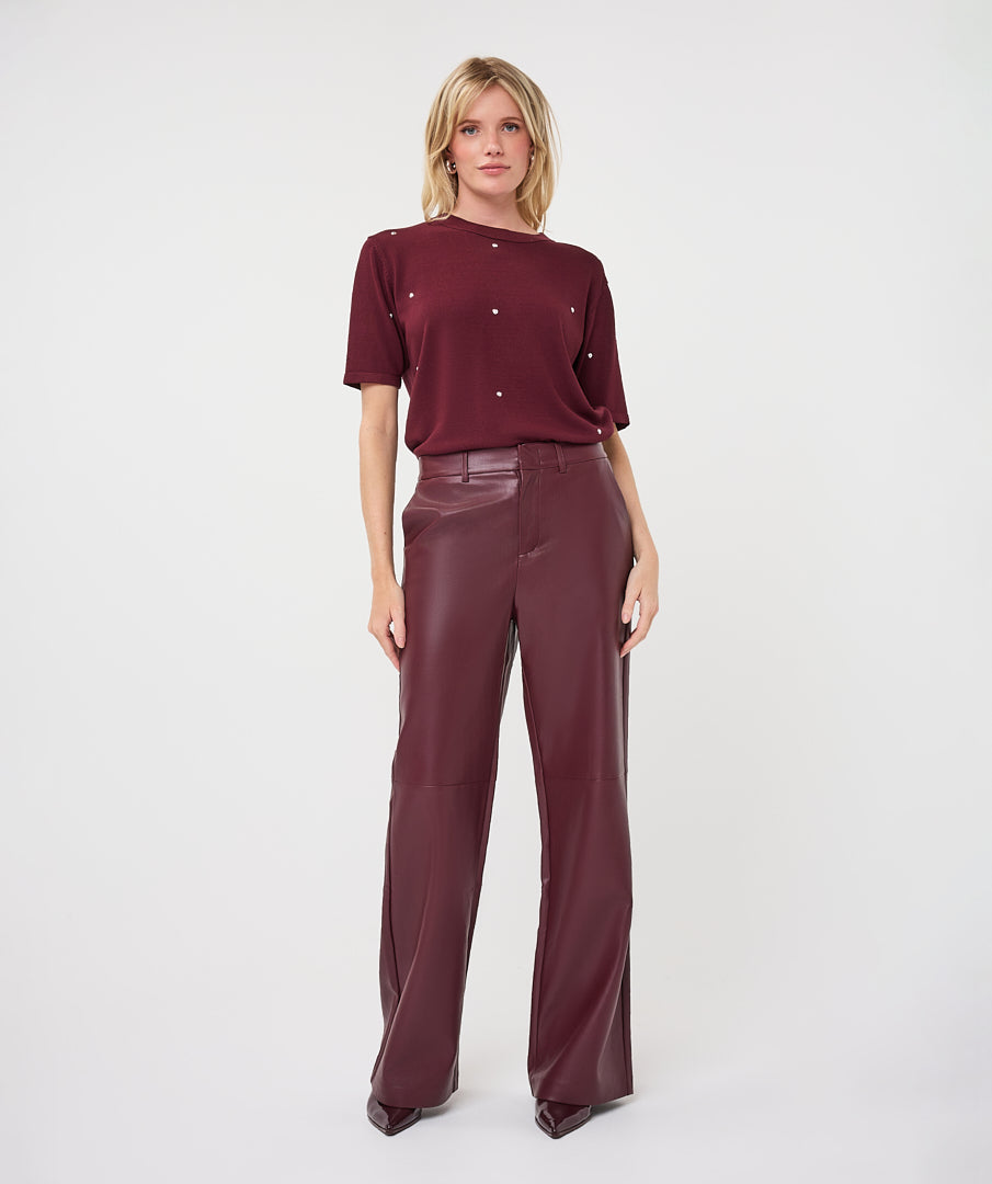 A model stands against a light gray studio background wearing a short-sleeve burgundy top with small metallic embellishments and matching wide-leg trousers.