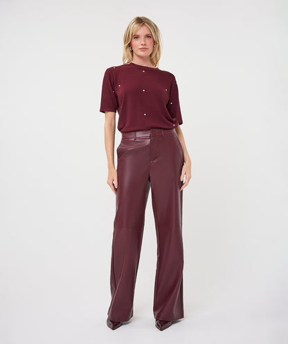 A model stands against a light gray studio background wearing a short-sleeve burgundy top with small metallic embellishments and matching wide-leg trousers.