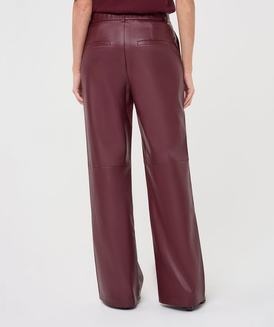 A person shown from the back wearing high-waisted, wide-leg trousers in a deep burgundy faux leather material, against a plain light gray studio background.