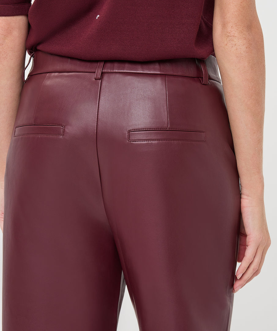 Close-up back view of a person wearing high-waisted burgundy faux-leather trousers with a matching top, against a light gray studio background.