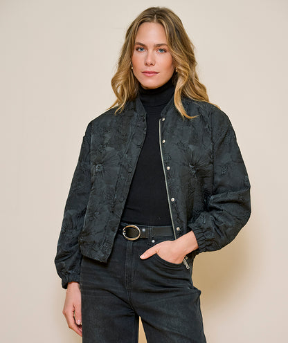 A woman modeling a dark charcoal gray textured bomber jacket with a floral jacquard pattern, worn over a black turtleneck and black jeans against a solid beige background.