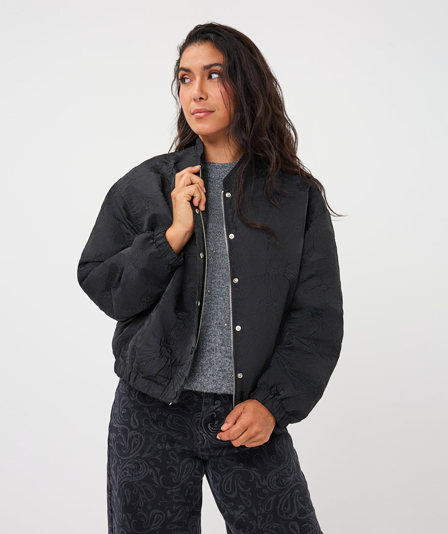 A woman modeling a black quilted bomber jacket with silver snap buttons, worn over a gray knit top and dark paisley-print pants, against a plain light gray studio background.