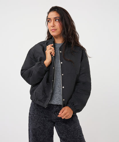 A woman modeling a black quilted bomber jacket with silver snap buttons, worn over a gray knit top and dark paisley-print pants, against a plain light gray studio background.