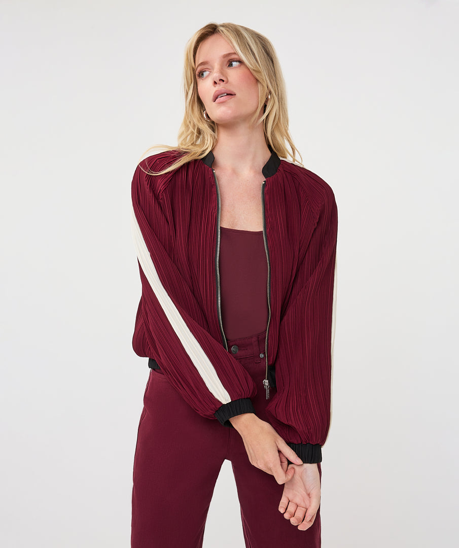 A woman modeling a pleated burgundy bomber jacket with cream diagonal stripes on the sleeves and black ribbed trim, paired with matching burgundy pants and a solid burgundy top against a light gray studio background.