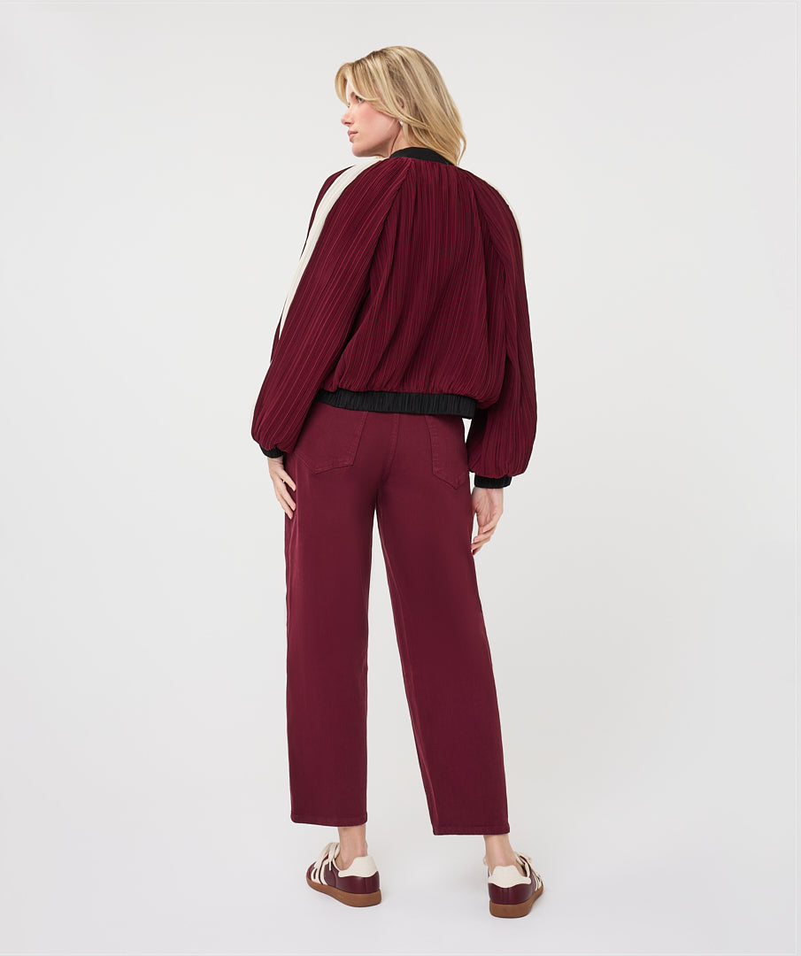 A woman shown from the back wearing a pleated burgundy bomber jacket with black ribbed trim, matching pleated trousers, and brown platform sandals, photographed against a clean white studio background.