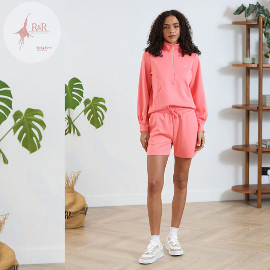 Woman wearing a pink tracksuit standing in a room with a plant and wooden shelf.
