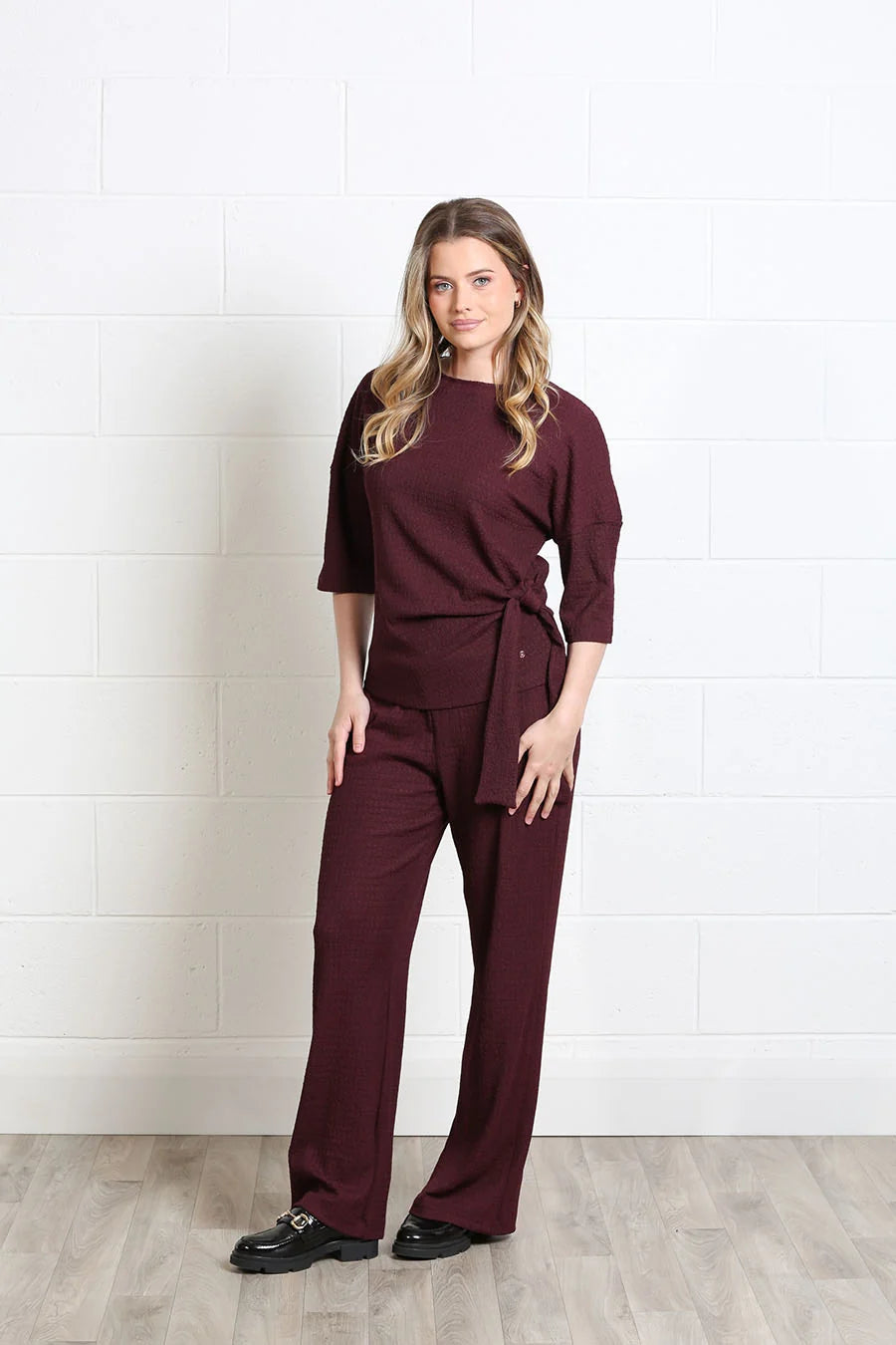 A woman modeling a matching burgundy wrap-style top and wide-leg pants set, standing against a white brick wall on a light wood floor.