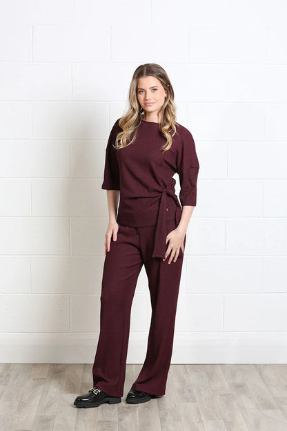 A woman modeling a matching burgundy wrap-style top and wide-leg pants set, standing against a white brick wall on a light wood floor.
