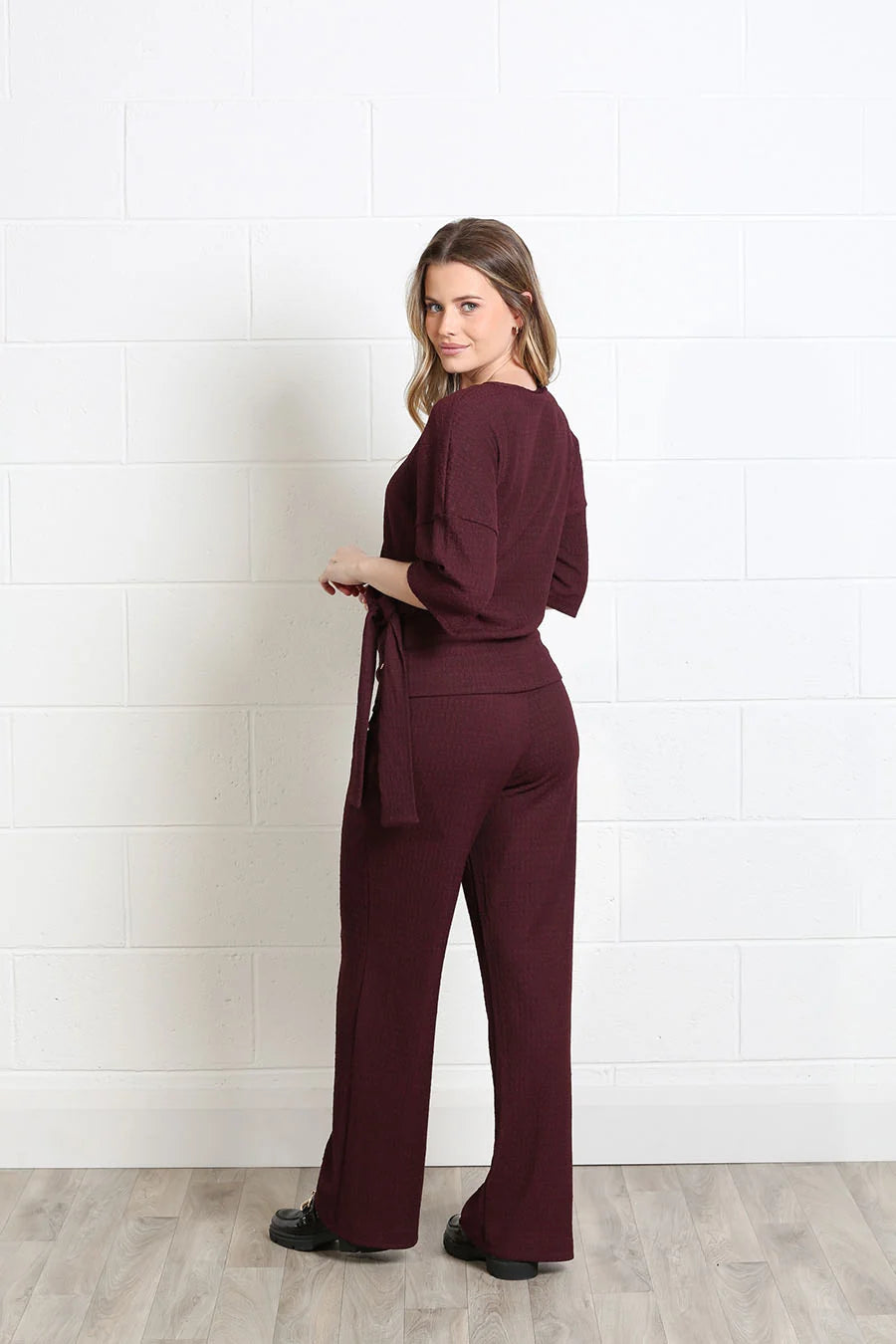 A woman modeling a burgundy three-piece jumpsuit with a tie waist, 3/4 sleeves, and wide-leg pants, standing against a white brick wall.