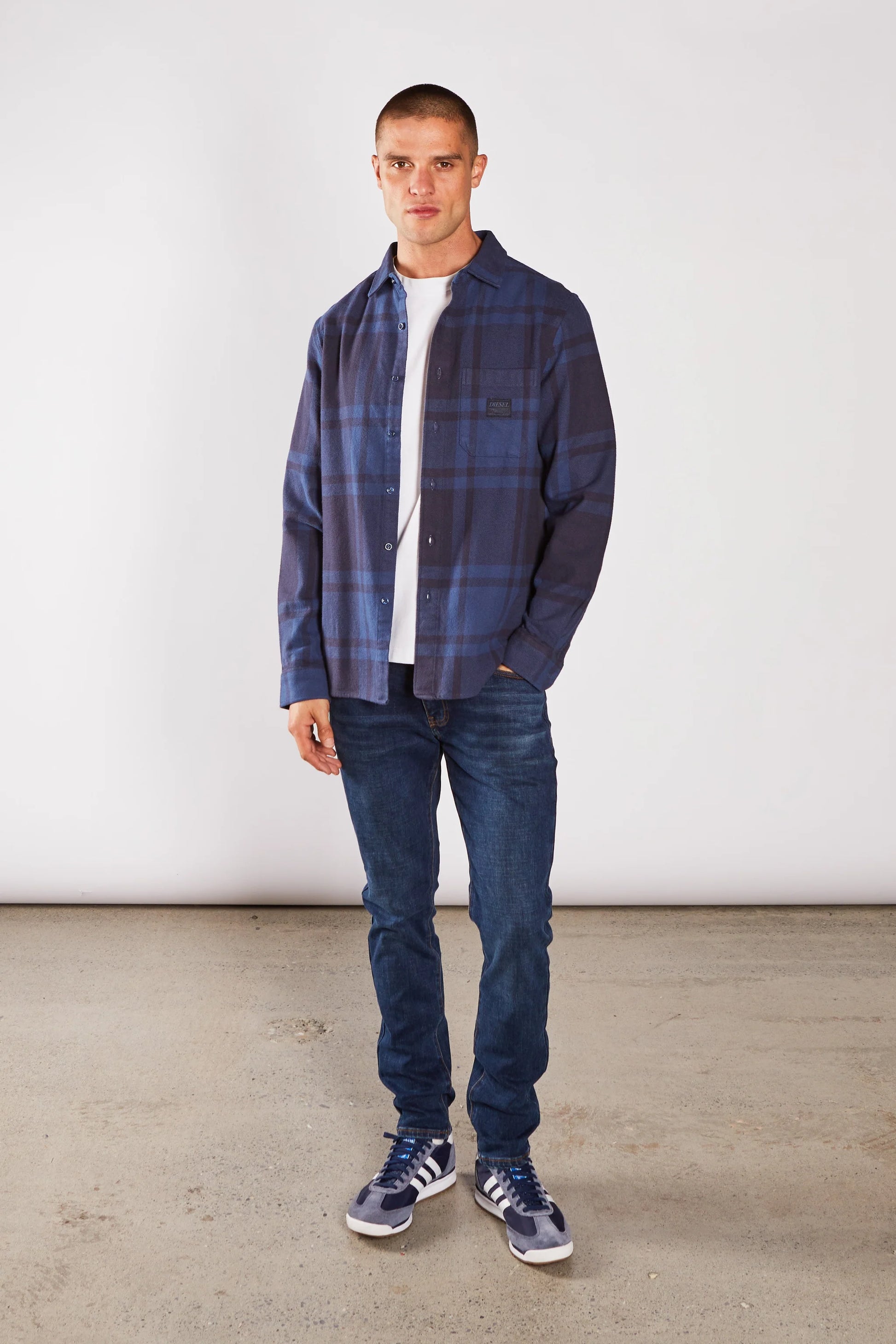 Man wearing a blue plaid shirt, white t-shirt, and jeans standing against a plain background