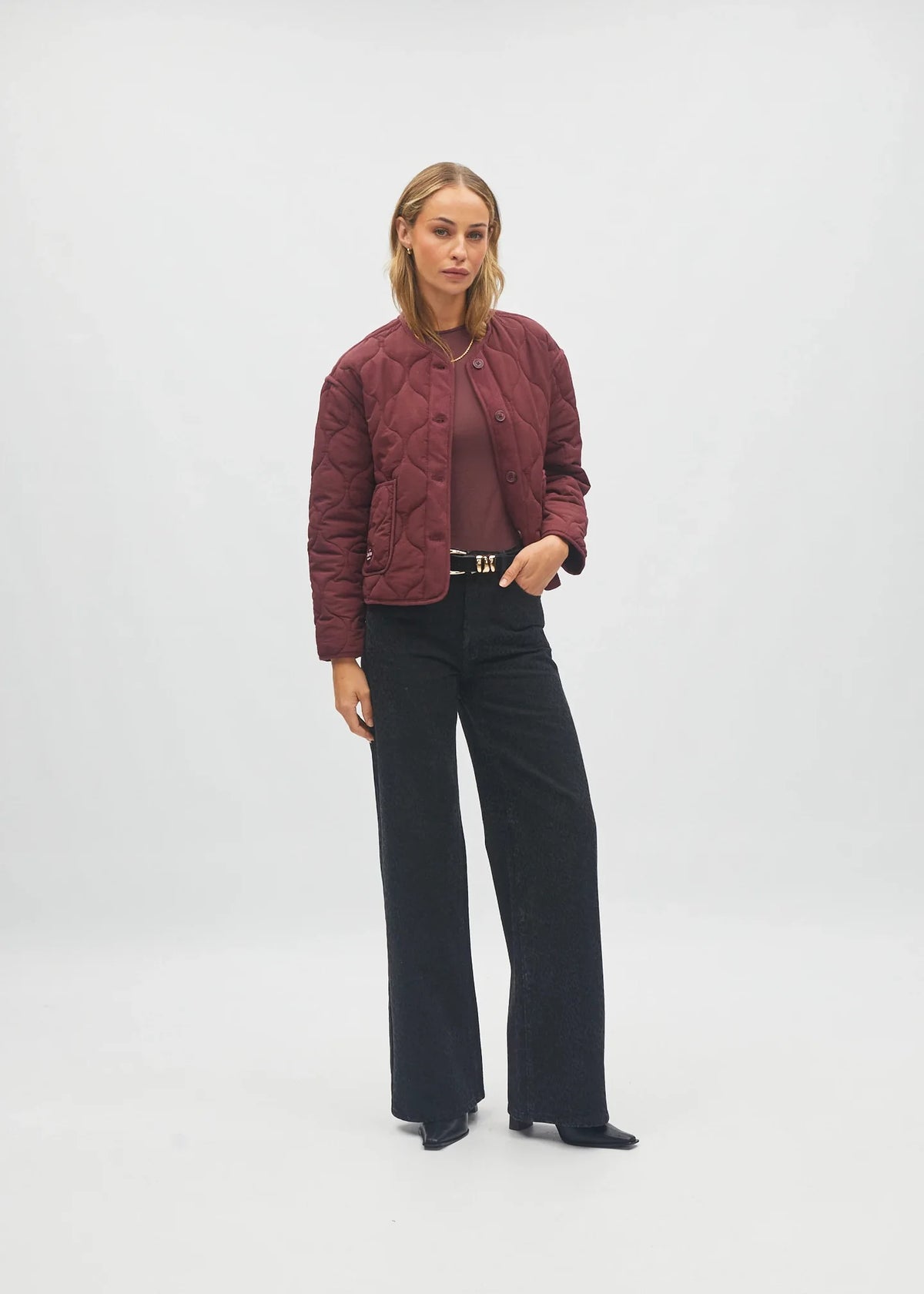 A model stands against a light gray studio background wearing a burgundy quilted cropped jacket over a matching burgundy top, black wide-leg trousers, and black pointed shoes.