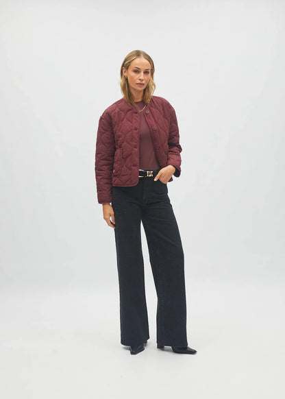 A model stands against a light gray studio background wearing a burgundy quilted cropped jacket over a matching burgundy top, black wide-leg trousers, and black pointed shoes.