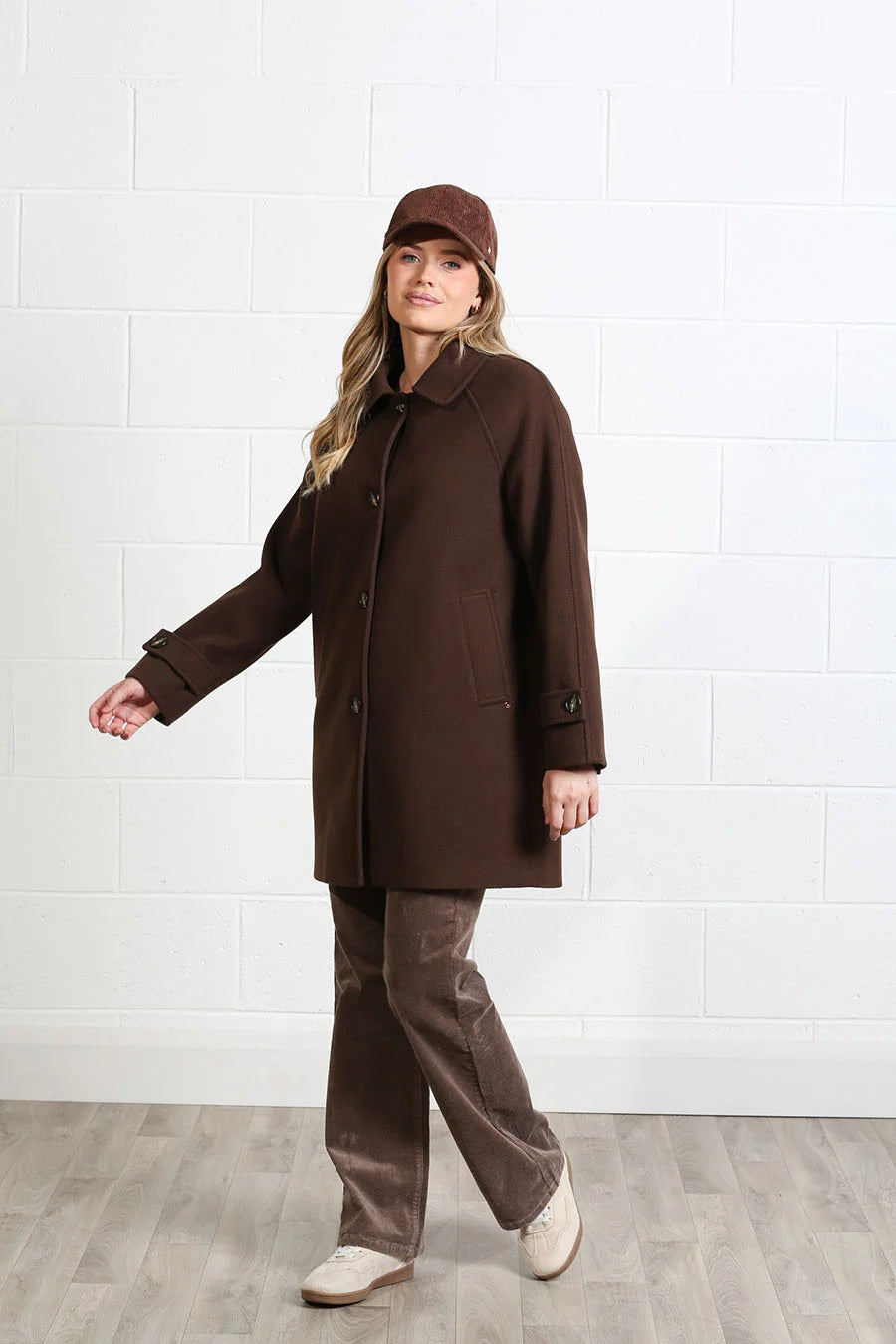 A woman modeling a dark brown wool coat, matching brown pants, a brown cap, and white shoes against a white brick wall background.