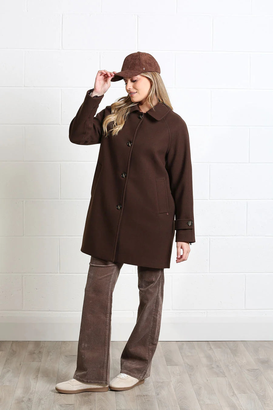 A woman modeling a dark brown wool-blend coat with a matching brown cap, brown corduroy pants, and beige slip-on shoes, standing against a white brick wall.