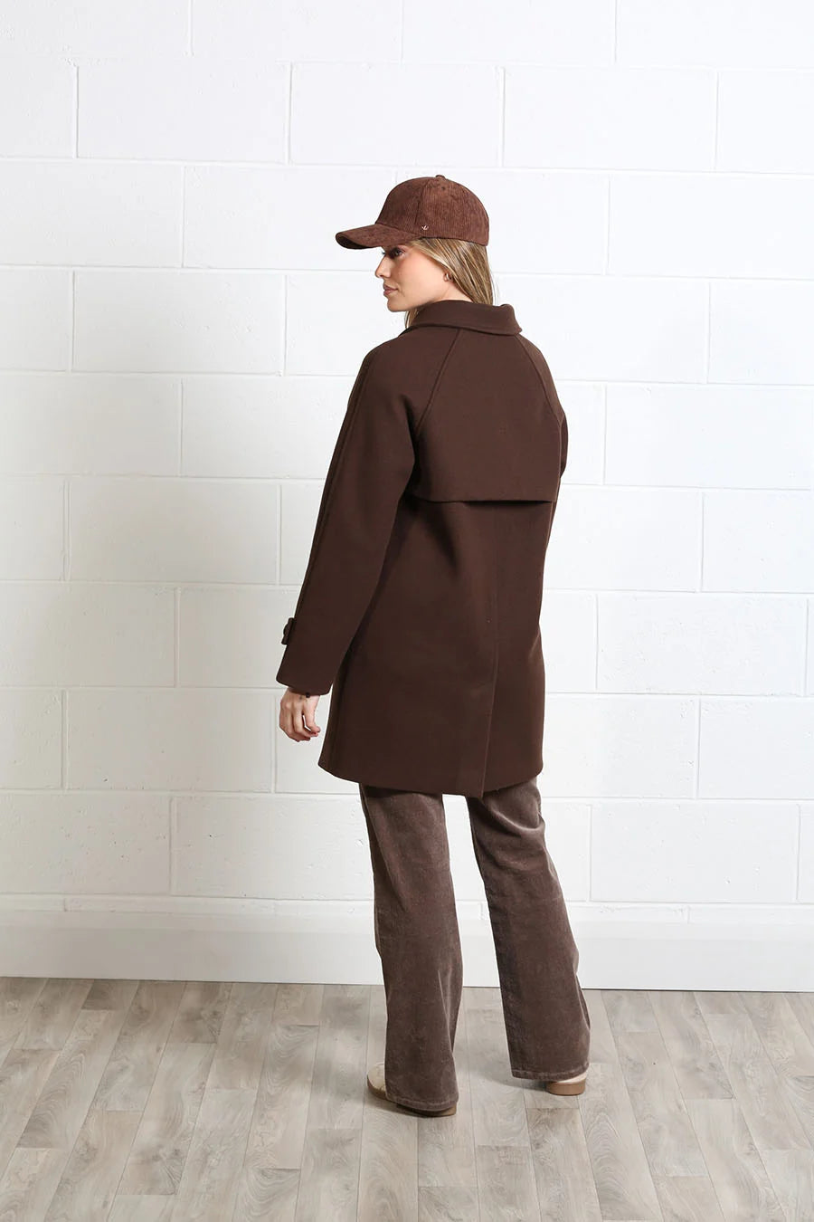 A model wearing a dark brown mid-length coat, matching brown pants, a brown cap, and brown shoes, standing in profile against a white brick wall and light wood floor.