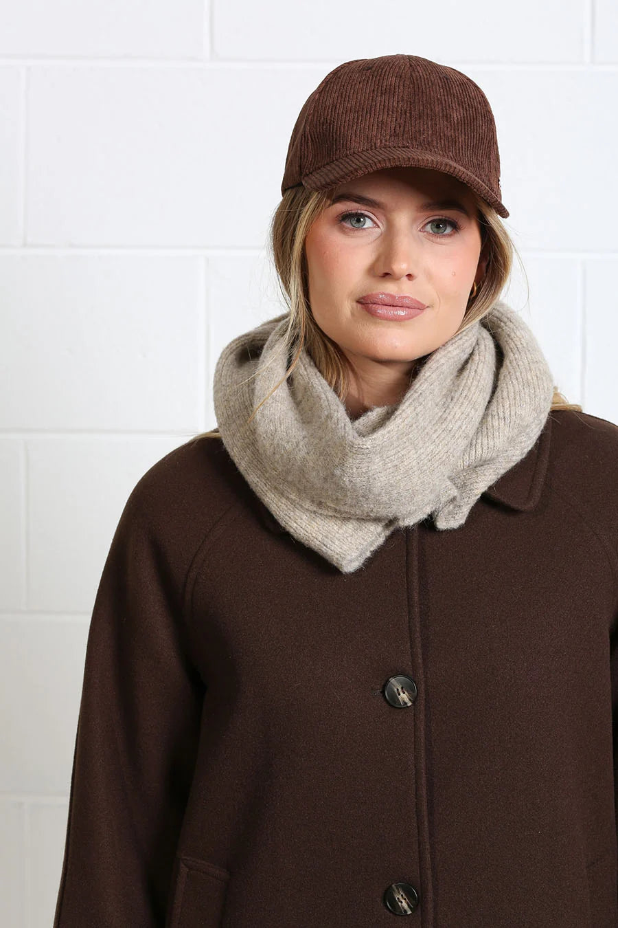 A woman wearing a brown corduroy cap, a thick beige knit scarf, and a brown wool coat, standing against a white tiled wall.