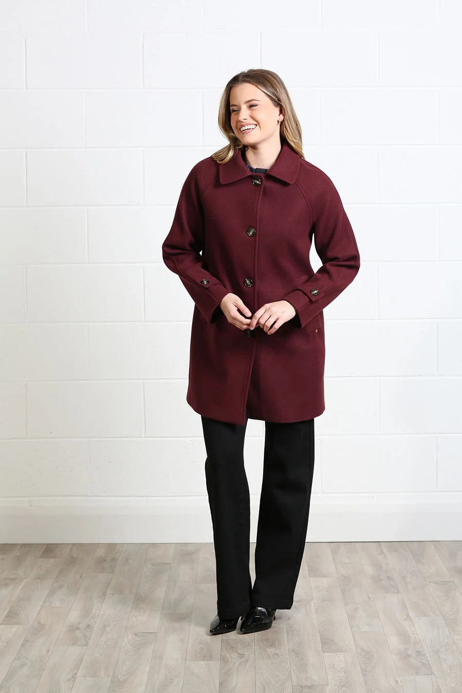 A woman modeling a burgundy double-breasted coat with black pants, standing against a white brick wall and light wood floor.