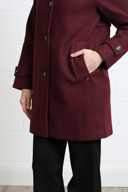 A person wearing a long burgundy wool-blend coat with dark buttons and a front patch pocket, paired with black trousers, standing against a white brick wall.