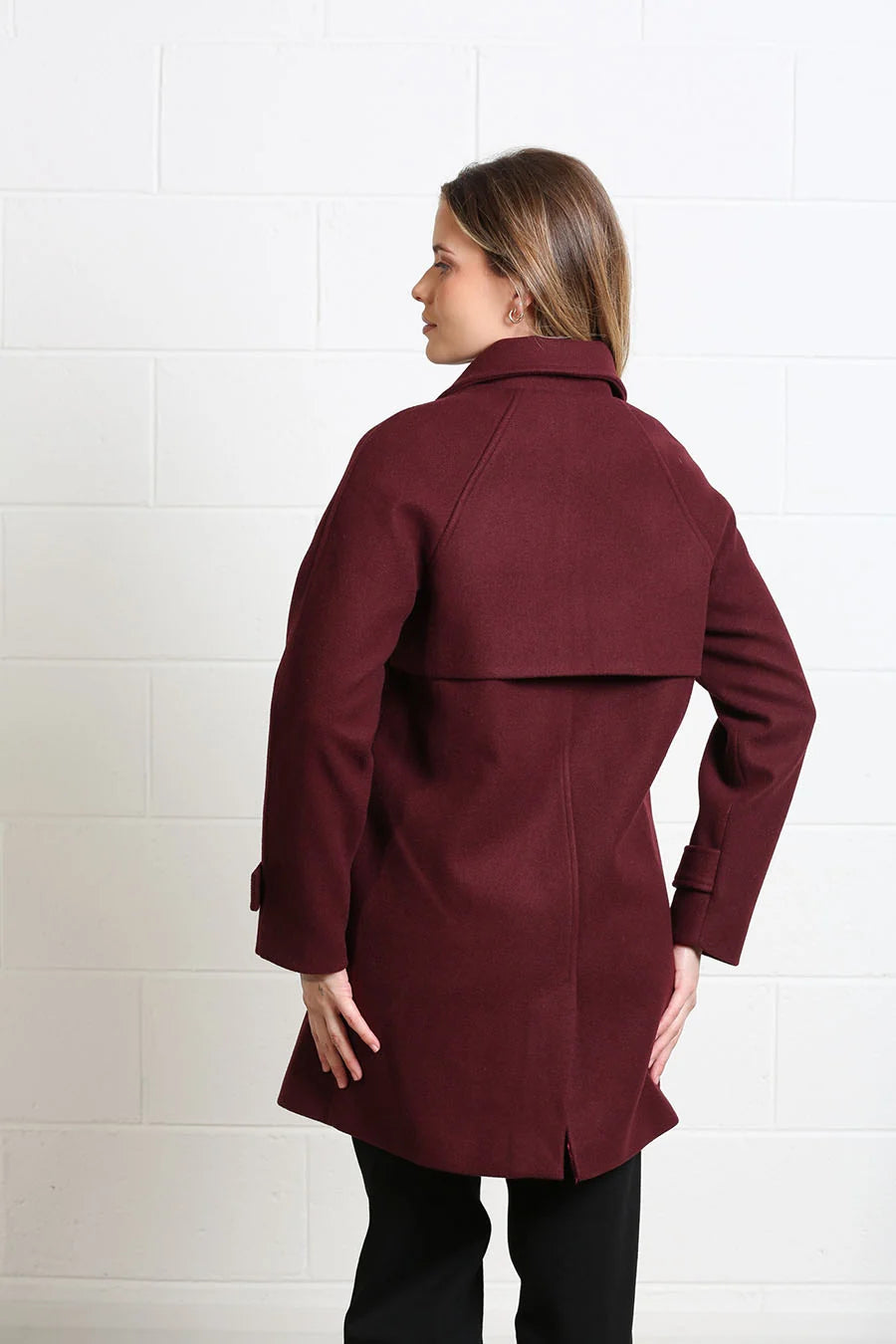 A woman wearing a mid-length maroon coat, shown from the back and side against a white brick wall.