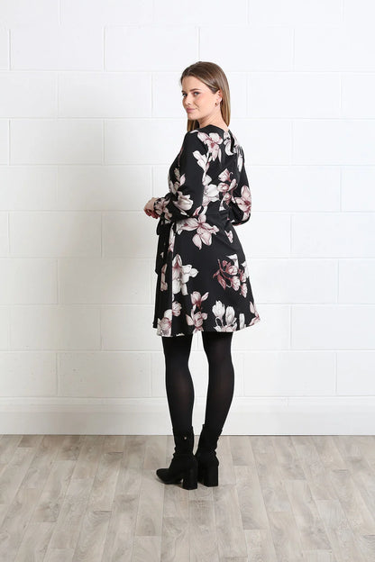 A woman modeling a black wrap-style mini dress with a white and pink floral pattern, paired with black tights and black ankle boots, standing against a white brick wall and light wood floor.