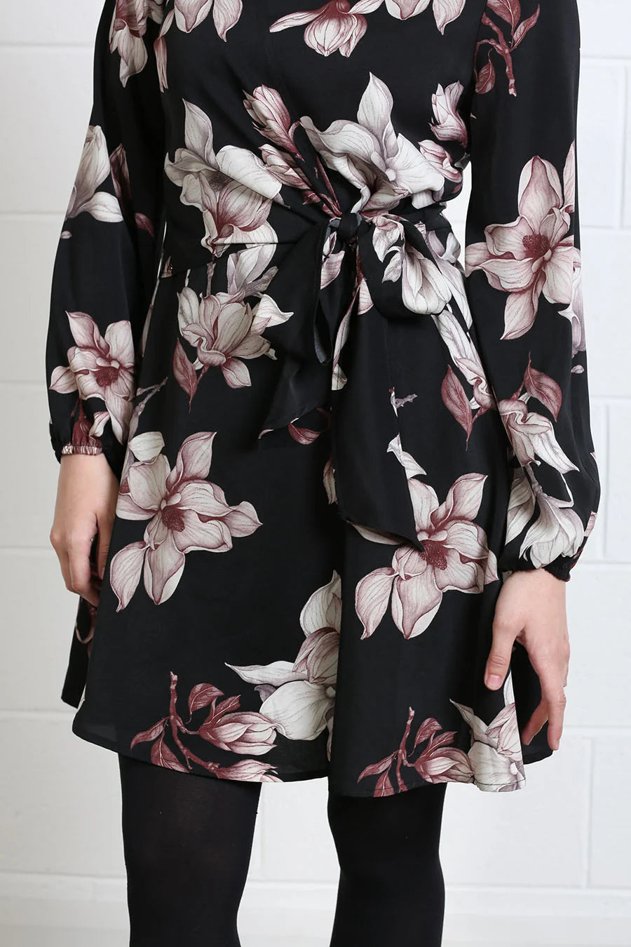A woman wearing a black long-sleeve wrap dress with a large floral print in dusty pink, mauve, and white. The dress features a tie waist and is paired with black tights, shown against a white brick wall background.