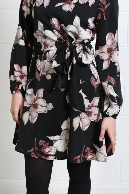 A woman wearing a black long-sleeve wrap dress with a large floral print in dusty pink, mauve, and white. The dress features a tie waist and is paired with black tights, shown against a white brick wall background.