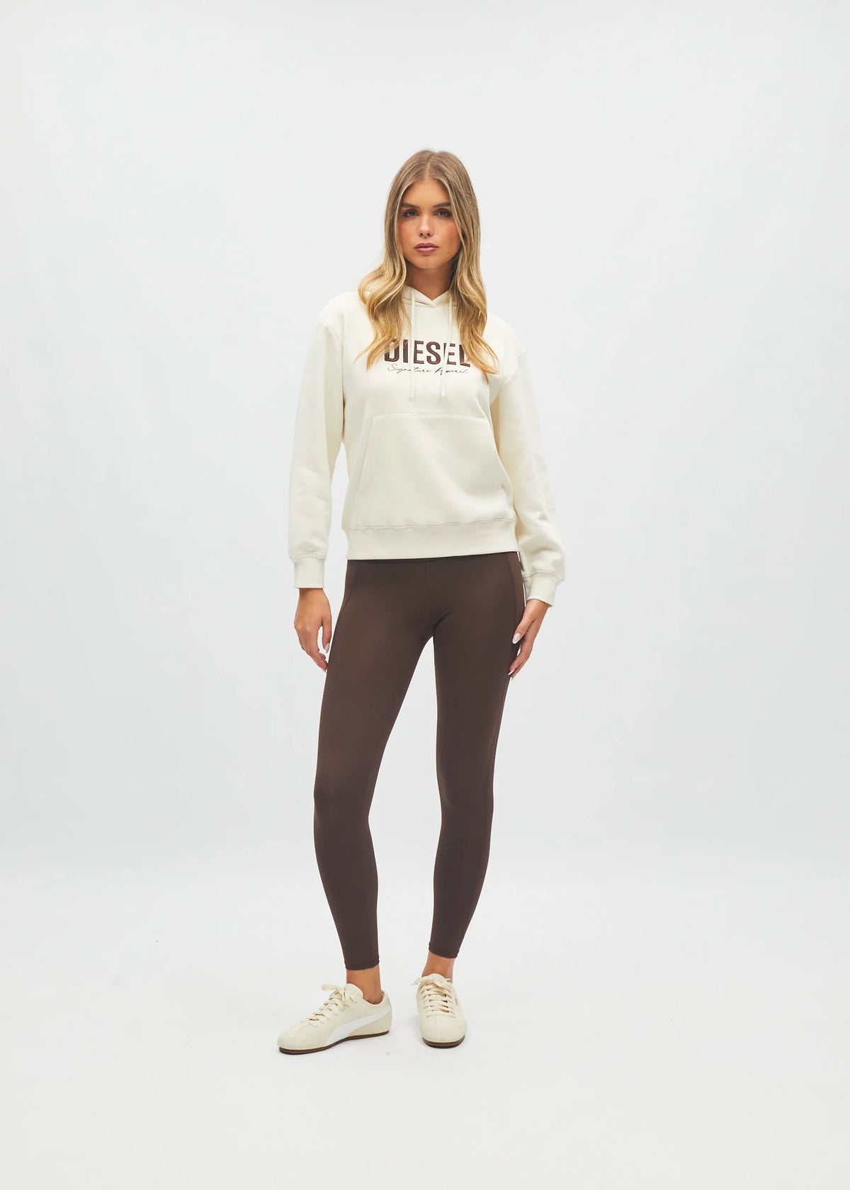Women modelling against plain background cream hoodie with logo and brown leggings