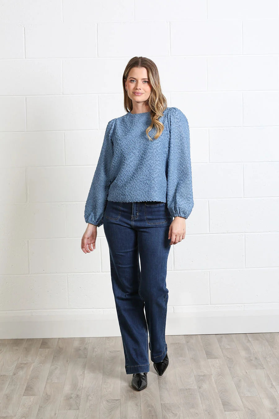 A woman modeling a textured blue long-sleeve sweater with puffed shoulders and dark blue jeans, standing against a white brick wall and light wood floor.