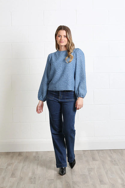 A woman modeling a textured blue long-sleeve sweater with puffed shoulders and dark blue jeans, standing against a white brick wall and light wood floor.