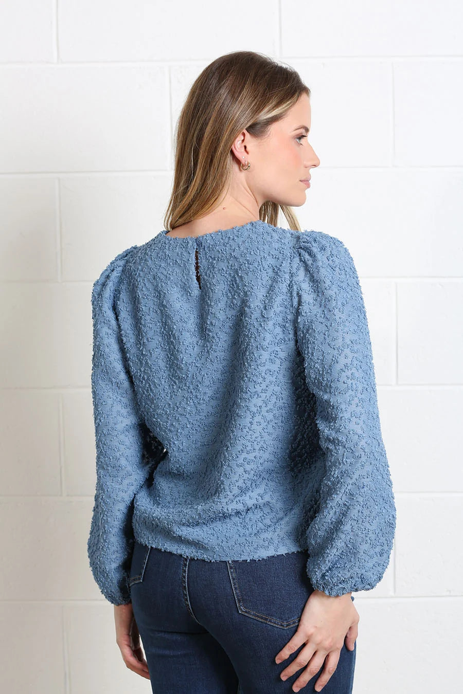 A woman shown from the back wearing a textured dusty blue long-sleeve top with puffed shoulders, paired with dark blue jeans, standing against a white brick wall.
