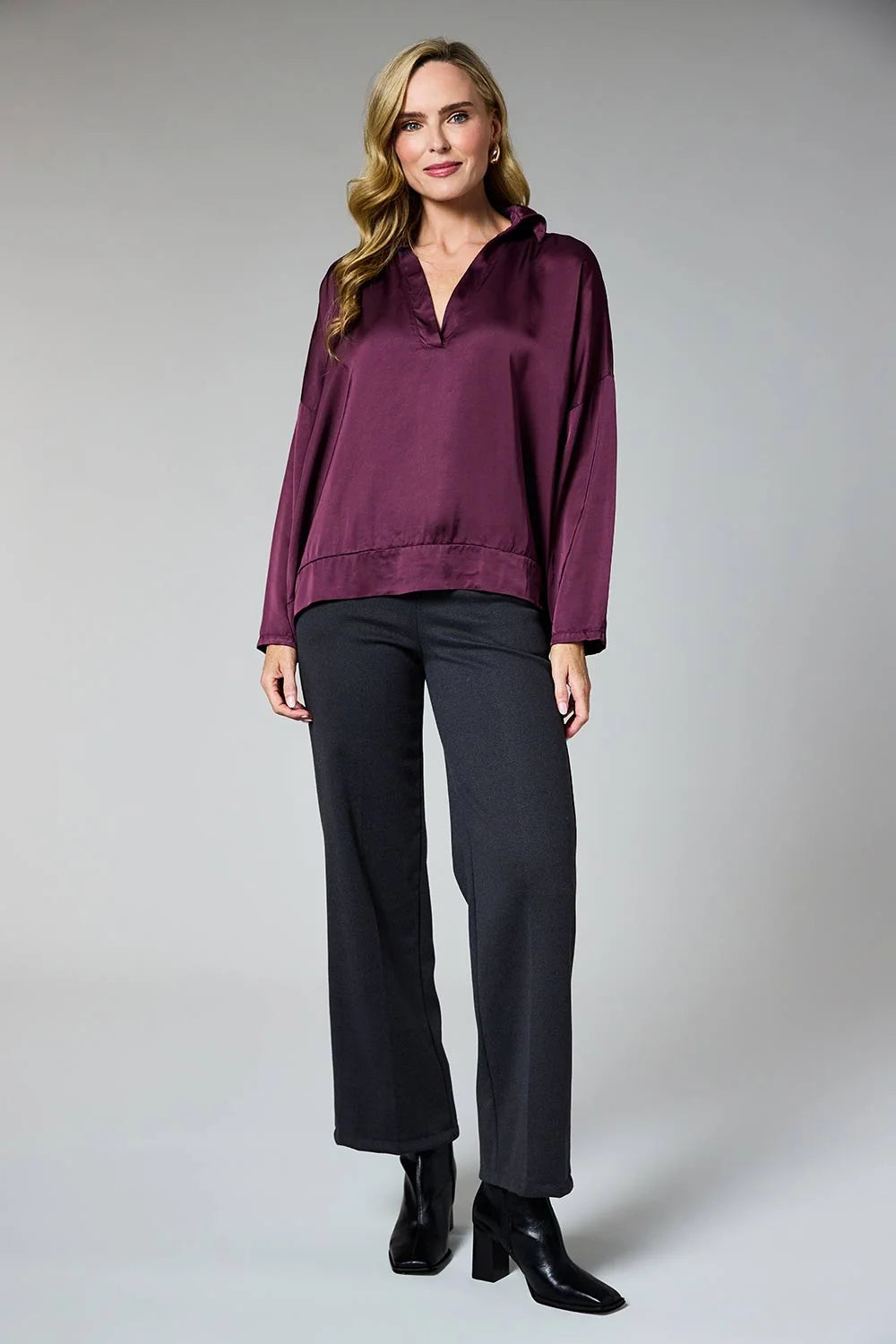 A woman modeling a long-sleeve satin half-placket blouse in deep plum purple, paired with dark charcoal wide-leg trousers and black ankle boots, against a neutral gray studio background.