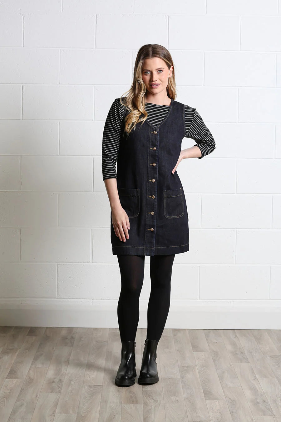 A woman modeling a dark navy denim pinafore dress over a black and white striped long-sleeve shirt, paired with black tights and black ankle boots, standing against a white brick wall.
