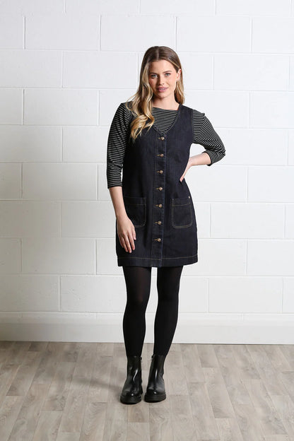 A woman modeling a dark navy denim pinafore dress over a black and white striped long-sleeve shirt, paired with black tights and black ankle boots, standing against a white brick wall.