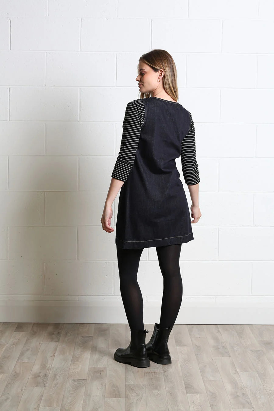 A woman stands in profile wearing a dark denim tunic dress with three-quarter black and white striped raglan sleeves, black tights, and black ankle boots, set against a white brick wall and light wood floor.