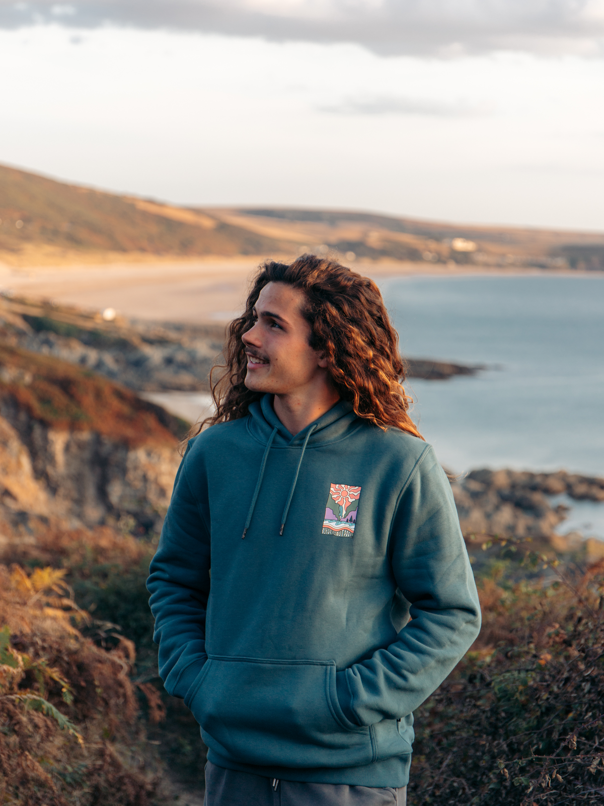 A man with long curly brown hair stands outdoors on a rocky coastal hillside, wearing a teal hoodie with a small colorful chest graphic. The background shows a beach, water, and hills under a cloudy sky.