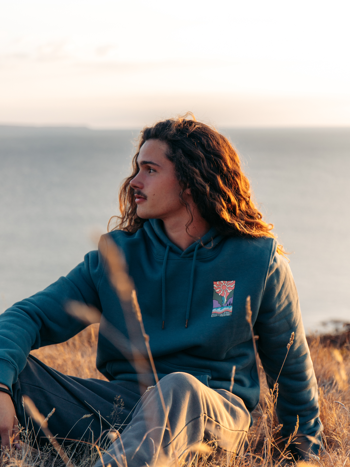 A man with long curly hair sits in dry grass outdoors by the ocean at sunset, wearing a teal hoodie with a small colorful chest graphic and light gray sweatpants.