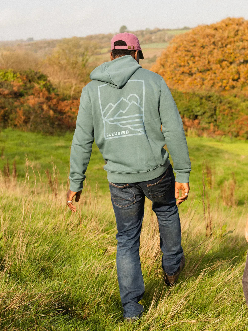 Person wearing a green hoodie with a mountain design walking through a field.