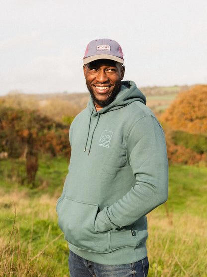 Man wearing a green hoodie and cap standing in a field with trees in the background