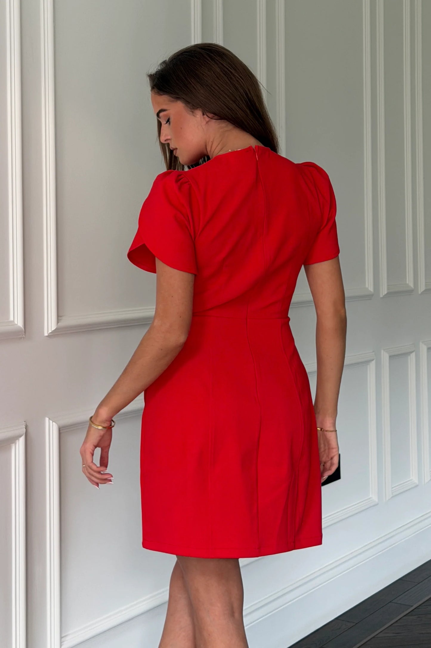 Woman wearing a red dress standing against a white paneled wall.