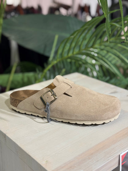 Beige clog shoe with a buckle on a wooden surface with green foliage in the background
