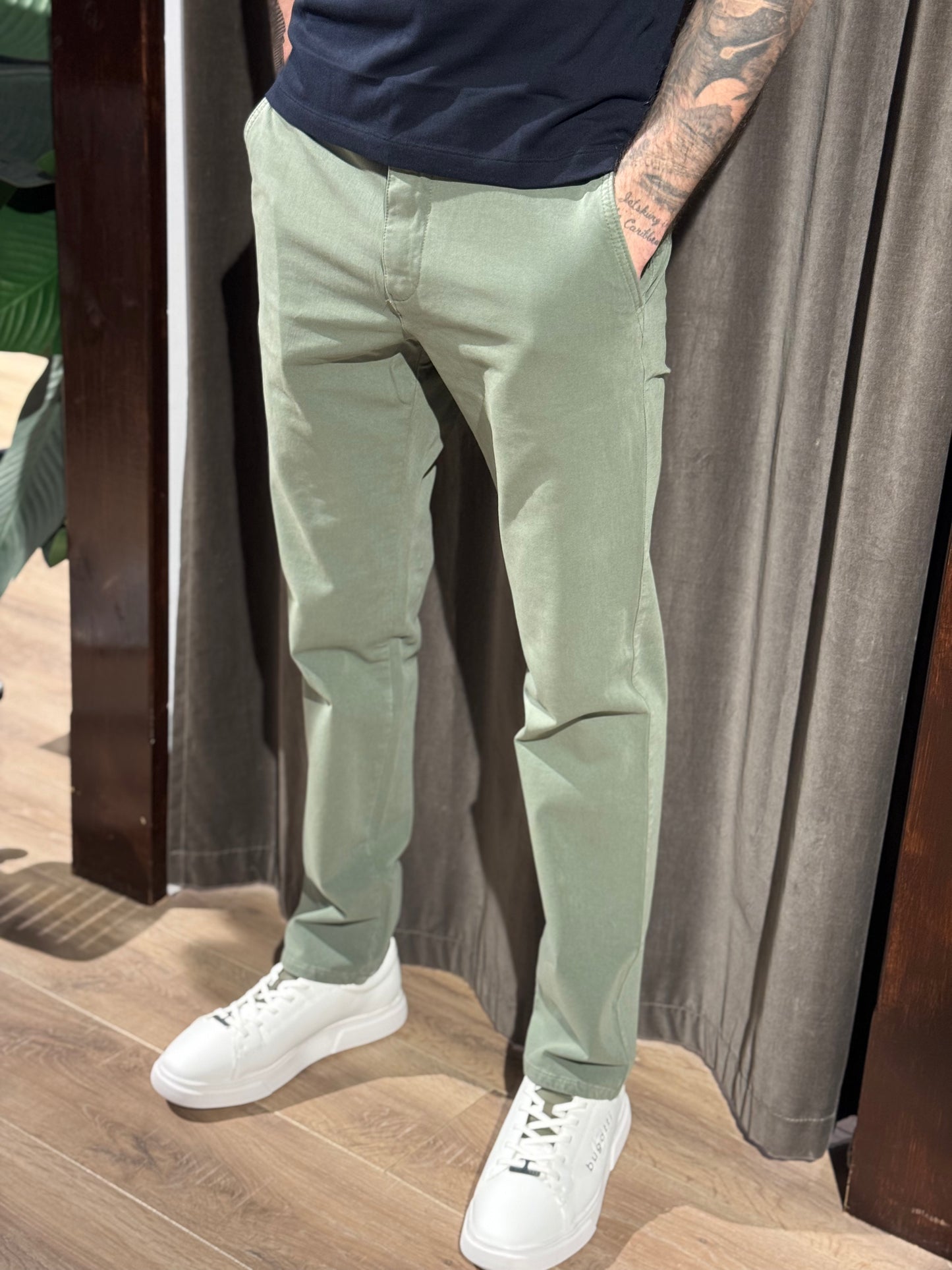 Person wearing light green pants and white sneakers standing indoors with a neutral background
