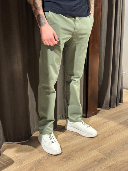 Person wearing green pants and white sneakers standing on a wooden floor with curtains in the background
