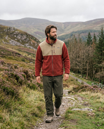 A man hiking on a grassy mountain path wearing a two-tone fleece jacket in rust red and beige, olive green trousers, and hiking boots, with mountains and trees in the background.