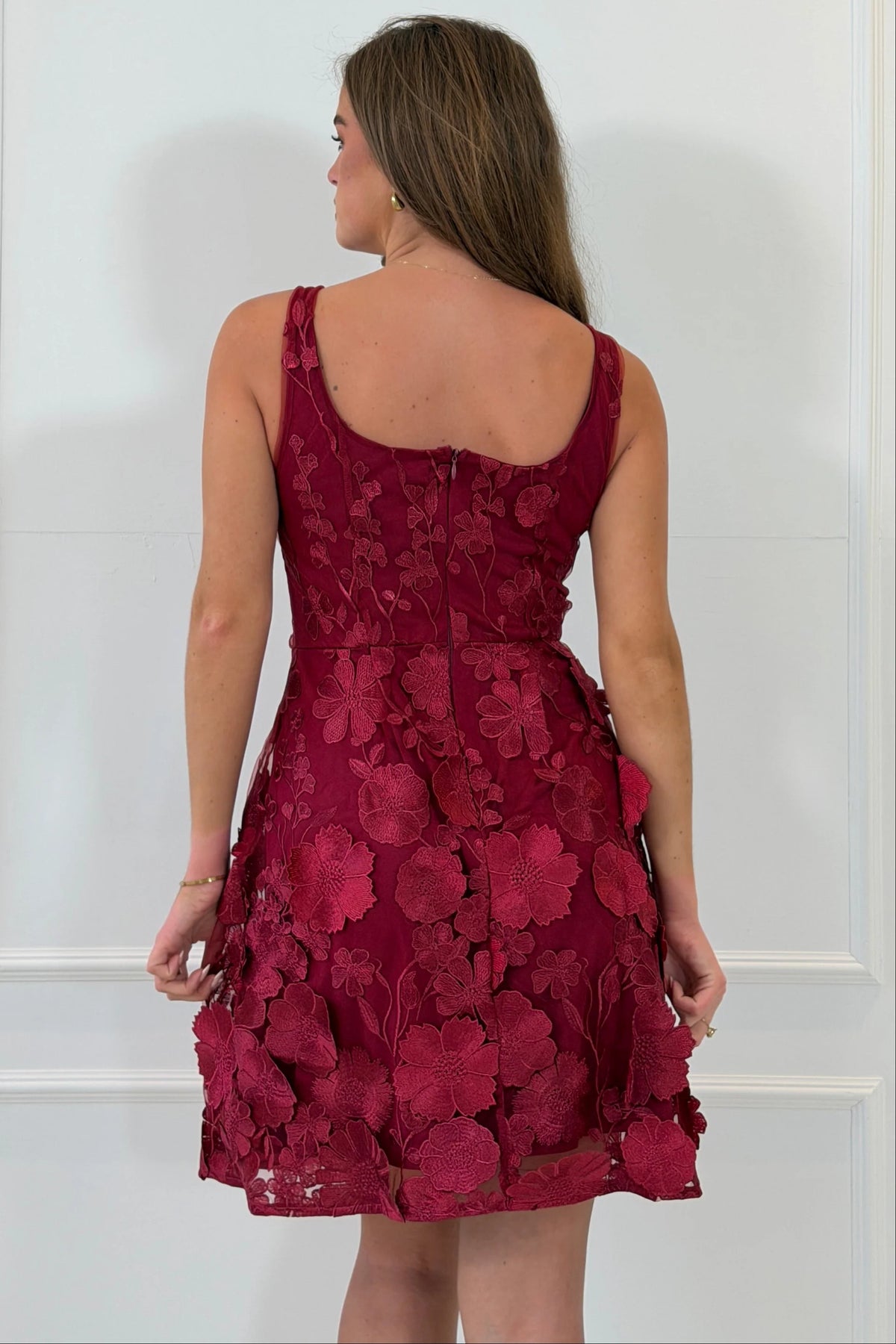 A woman shown from the back wearing a sleeveless burgundy dress with a floral lace overlay and a fitted bodice, photographed against a white paneled wall.
