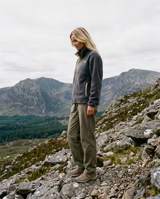 A woman stands on a rocky mountain slope wearing a dark gray quarter-zip fleece jacket, olive green hiking pants, and brown hiking boots, with mountains and a cloudy sky in the background.